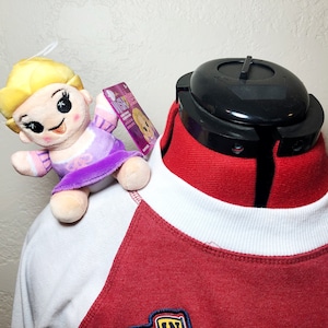 Könnte beinhalten: Ein weiß-rotes Baseball-Shirt mit weißem Kragen. Auf der Schulter des Shirts sitzt ein kleines Plüschtier einer Prinzessin mit blonden Haaren und einem lila Kleid.