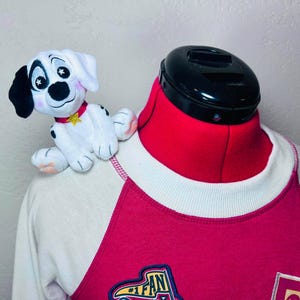 May include: A plush dalmatian puppy toy with black ears and star-shaped pupils, wearing a red collar with a yellow star charm, perched on the shoulder of a red and white raglan-style sweatshirt featuring a #1 FAN patch on the front.