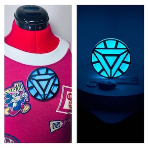 Op de afbeelding: Een cirkelvormig arc-reactor rekwisiet met een zwart frame en een oplichtend lichtblauw driehoekig centrum. Het object is bevestigd aan een rood kledingstuk met Mickey Mouse-patches en wordt ook losstaand en verlicht getoond.