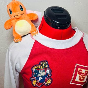 May include: Red and white baseball style long-sleeved shirt with a Mickey Mouse graphic and the text "#1 FAN". The shirt also has a red, yellow, and white patch with the text "1971" and a Mickey Mouse head graphic.
