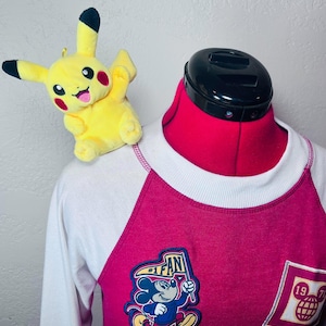 Puede incluir: Una camiseta raglán blanca y magenta con un parche gráfico de Mickey Mouse sosteniendo un banderín que dice #1 FAN y un parche cuadrado con el texto 1971 y un icono de globo terráqueo. Un pequeño peluche amarillo de Pikachu descansa sobre el hombro.