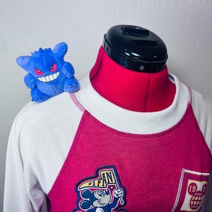 May include: A white and magenta raglan sweatshirt featuring a Mickey Mouse patch with the text #1 FAN and a square patch with the number 1971. A small blue Gengar plush toy with red eyes and a wide white grin is perched on the shoulder of the sweatshirt.