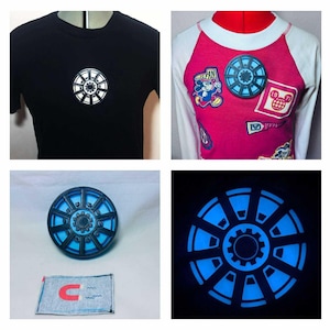 Może przedstawiać: Kolaż zdjęć przedstawiających projekty reaktorów łukowych Iron Mana. Jedno zdjęcie przedstawia czarny t-shirt z białym i srebrnym emblematem reaktora. Inne przedstawia różowo-białą koszulkę raglan z dwiema naszywkami reaktora. Dodatkowe zdjęcia pokazują reaktor 3D i świecący na niebiesko reaktor.