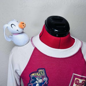 May include: A raglan sleeve sweatshirt with a white body and magenta sleeves. The sweatshirt features a Mickey Mouse patch that says "#1 FAN" and a small plush snowman with a carrot nose. The sweatshirt is displayed on a red dress form.