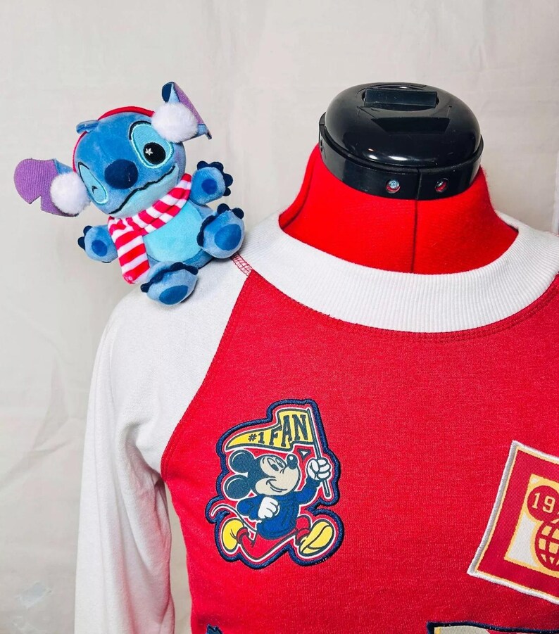 May include: Red and white long-sleeved shirt with a Mickey Mouse graphic that says "#1 FAN" and a blue and white Stitch plush toy on the shoulder.