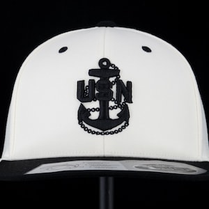 May include: A white baseball cap with a black brim and a black embroidered anchor logo on the front. The anchor features the letters USN integrated into its design, with a chain detail wrapping around the anchor base. A sticker is on the brim.