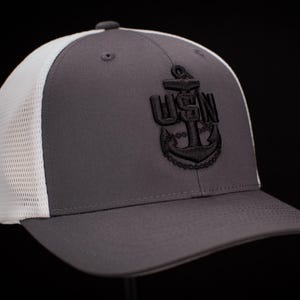 May include: A gray and white trucker hat with a black embroidered anchor and the text "USN".
