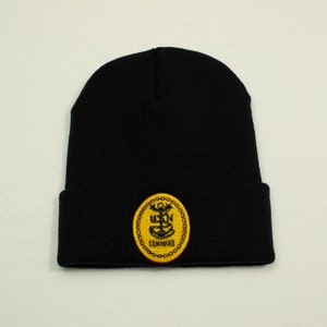 May include: Black knit beanie with a folded brim. A yellow and black embroidered patch with the text "USN COMMAND" and an anchor design is sewn on the front. The beanie is made of a soft, knitted material.