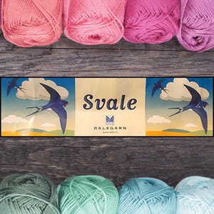 Puede incluir: Una colección de ovillos de lana coloridos en tonos rosa, verde y azul, dispuestos sobre una superficie de madera. En el centro hay una caja con la palabra "Svale" y una ilustración de golondrina. La lana es probablemente para tejer o hacer ganchillo.