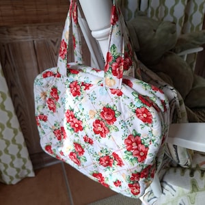 Puede incluir: Un bolso blanco acolchado con un estampado floral de rosas rojas y hojas verdes. El bolso tiene forma cuadrada con cierre de cremallera y dos asas. El bolso está colgado de una silla blanca.