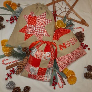 May include: Three beige drawstring gift bags with festive red and white patchwork designs. One bag features a star, another has "NOEL" appliqued, and the third has a patchwork pattern. The bags are decorated with candy canes, pine cones, and dried orange slices.