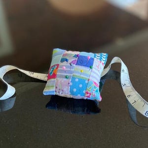 May include: A patchwork pincushion with various fabric patterns and colors, including blue polka dots and floral prints. Several pins with colorful heads are inserted into the cushion. A white measuring tape is partially visible.