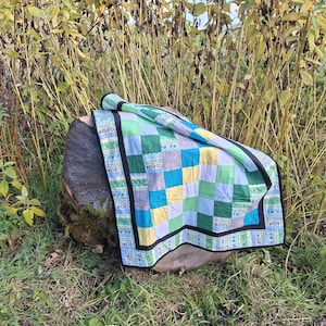 May include: A patchwork quilt featuring a grid of squares in shades of green, blue, yellow, and white. The quilt is bordered by a black fabric trim and a patterned strip with small green leaf motifs, finished with a solid black edge binding.