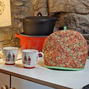 Op de afbeelding: Een feestelijke keukenscène met een rood-groene bloemen theemuts, twee witte mokken met kerstontwerpen en een zwarte gietijzeren pot bovenop een rode pot. De theemuts heeft een groene rand.