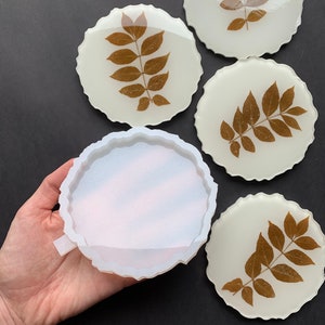 Puede incluir: Conjunto de cuatro posavasos de resina blanca con un diseño de hojas marrones. Los posavasos tienen un borde festoneado y están hechos con una resina transparente que muestra las hojas en el interior. También se muestra un molde de silicona para hacer los posavasos.