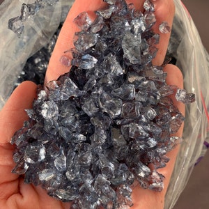 May include: A close-up of a handful of iridescent, clear glass pieces with a slight blue tint. The glass pieces are irregular in shape and size, and they are scattered in a pile.