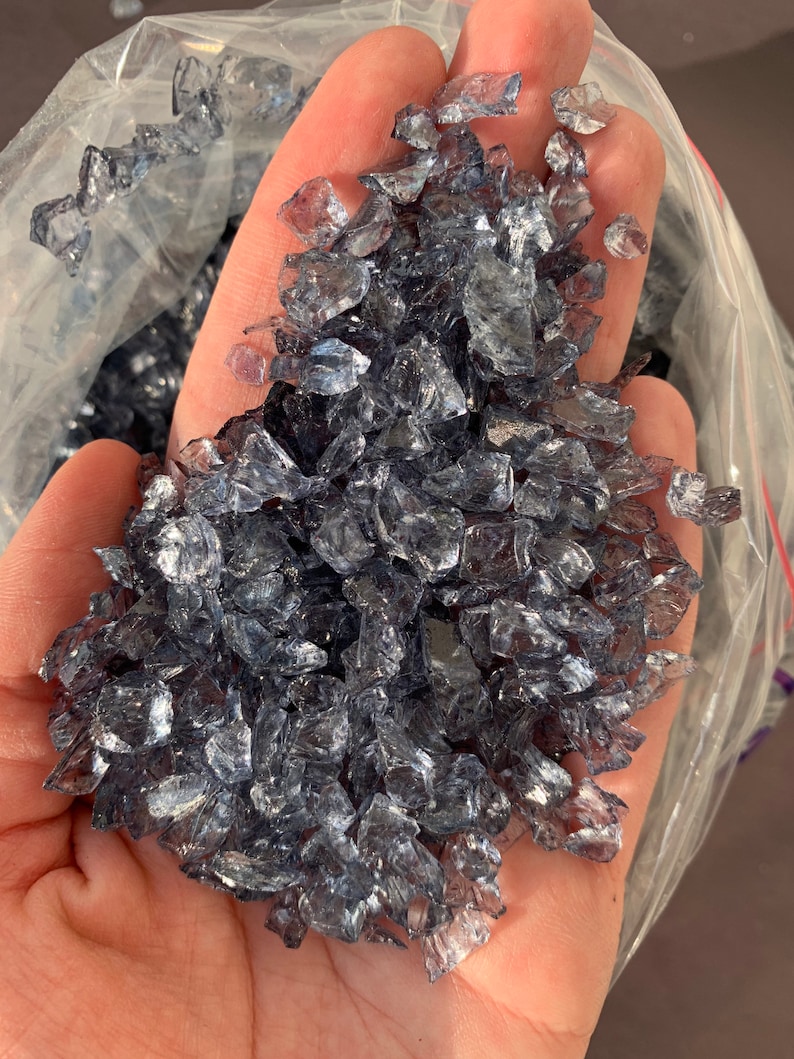 May include: A close-up of a handful of iridescent, clear glass pieces with a slight blue tint. The glass pieces are irregular in shape and size, and they are scattered in a pile.