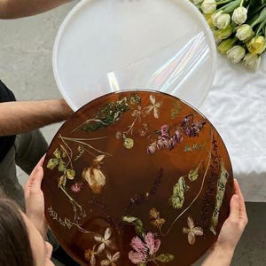 Puede incluir: Una bandeja redonda de resina color ámbar con flores y vegetación secas incrustadas. La bandeja se sostiene frente a una tapa o base circular blanca translúcida, mostrando un acabado liso y brillante con detalles botánicos visibles.