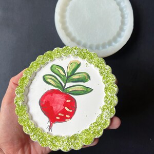Può includere: Un sottobicchiere rotondo in resina bianca con un bordo di glitter verde e un ravanello rosso dipinto a mano con foglie verdi.