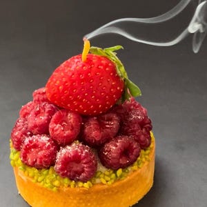 May include: A small pastry tart with a golden crust topped with fresh raspberries and a single strawberry. The tart is on a dark gray surface with a wisp of smoke rising from the top.