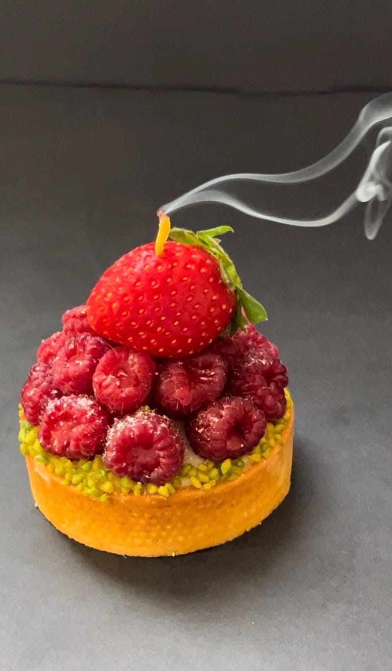 May include: A small pastry tart with a golden crust topped with fresh raspberries and a single strawberry. The tart is on a dark gray surface with a wisp of smoke rising from the top.