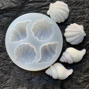 Puede incluir: Un molde de silicona con cuatro formas de conchas marinas y cinco conchas marinas blancas con purpurina iridiscente. Las conchas marinas están hechas de resina o arcilla polimérica.