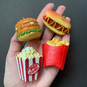 Puede incluir: Cuatro adornos de comida en miniatura: una hamburguesa, un perrito caliente, papas fritas en una caja roja y palomitas de maíz en una caja a rayas rojas y blancas con la palabra "POP CORN" en la parte delantera.