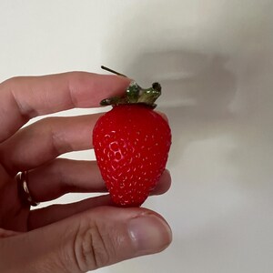 May include: A single, bright red strawberry with a green leafy top. The strawberry is held in a hand.