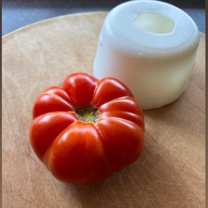 May include: A bright red tomato with a rounded shape sits on a wooden chopping board next to a white ceramic bowl.