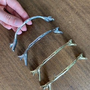 May include: Three decorative cabinet pulls in the shape of a branch. The pulls are made of metal and are available in silver and gold finishes.