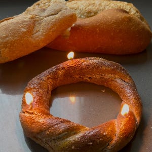 Op de afbeelding: Een bruine broodring met drie brandende kaarsen erin. De ring ligt op een bruin oppervlak. Twee broden zijn op de achtergrond.