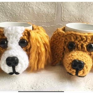 Op de afbeelding: Twee gehaakte hondenbekerwarmers. Eén is wit en bruin, de andere is bruin. Beide hebben zwarte ogen en neuzen.