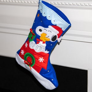 May include: Blue and red Christmas stocking with a white felt cuff. The stocking features an embroidered image of Snoopy in his doghouse.