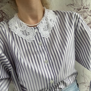 Pode incluir: Uma camisa de manga comprida listrada com uma gola de renda branca. A camisa &eacute; de cor roxo claro e branco. A camisa est&aacute; enfiada em uma cal&ccedil;a jeans azul claro.