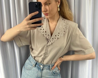 May include: A woman wearing a beige blouse with a white lace collar and light blue jeans. She is holding a black smartphone in her right hand and has her left hand on her hip.