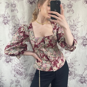 May include: A woman wearing a red and floral print blouse with a square neckline and puffed sleeves. She is also wearing black pants with a gold chain belt.