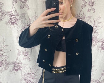 Puede incluir: Una chaqueta corta de terciopelo negro con botones dorados y un cinturón de cadena dorada. La chaqueta tiene un escote redondo y mangas largas.