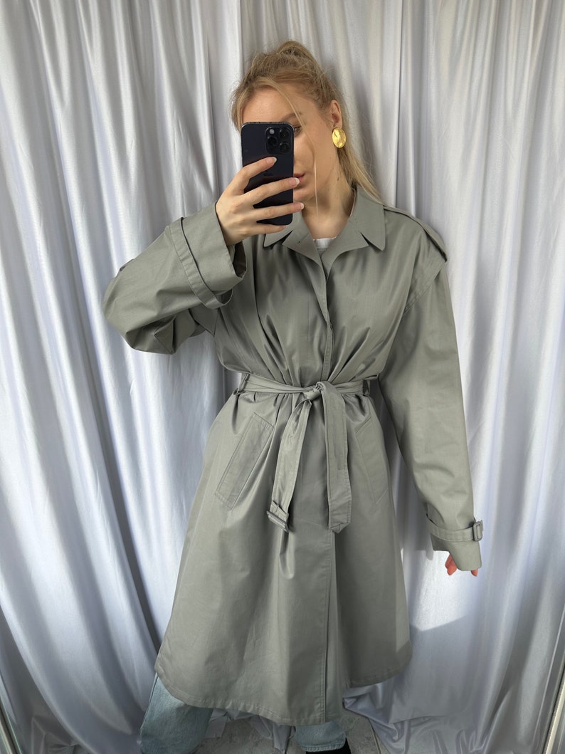 May include: A gray trench coat with a belt and long sleeves. The coat has a button closure and a collar.