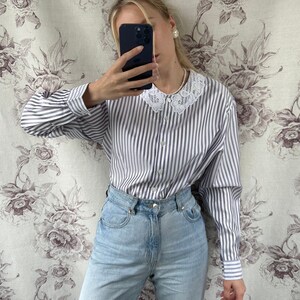 Pode incluir: Uma camisa de manga comprida listrada em branco e azul com uma gola de renda branca. A camisa tem um fecho de bot&otilde;es na frente e &eacute; usada com cal&ccedil;as de ganga azul claro.