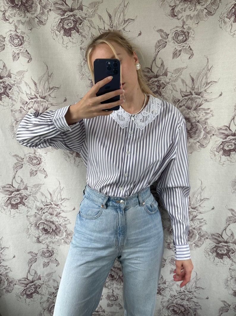 Pode incluir: Uma camisa de manga comprida listrada em branco e azul com uma gola de renda branca. A camisa tem um fecho de bot&otilde;es na frente e &eacute; usada com cal&ccedil;as de ganga azul claro.