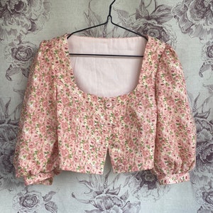 May include: A pink floral print cropped blouse with puffed sleeves and a square neckline. The blouse is hanging on a black hanger against a floral patterned background.