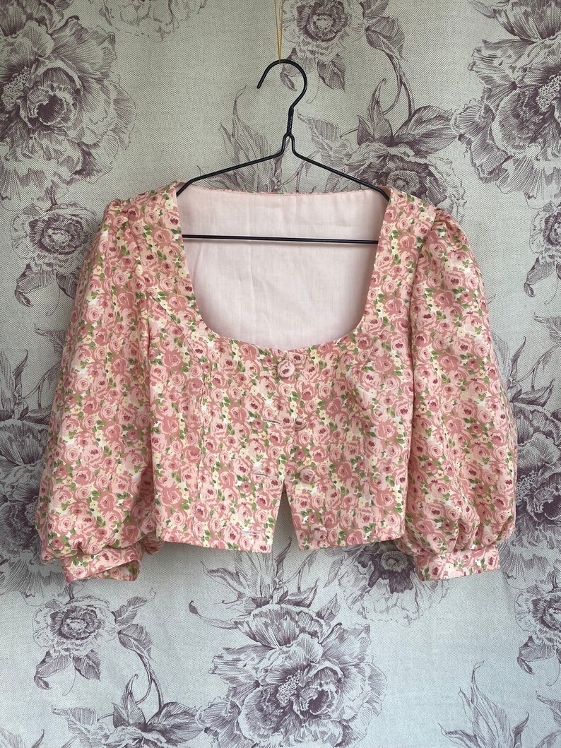 May include: A pink floral print cropped blouse with puffed sleeves and a square neckline. The blouse is hanging on a black hanger against a floral patterned background.