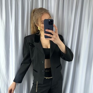 Może przedstawiać: Czarny blazer z aksamitnym kołnierzem i złotym łańcuszkowym paskiem. Blazer jest kr&oacute;tki i zapinany na jeden guzik. Modelka ma na sobie czarne spodnie.