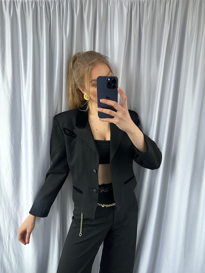 Może przedstawiać: Czarny blazer z aksamitnym kołnierzem i złotym łańcuszkowym paskiem. Blazer jest kr&oacute;tki i zapinany na jeden guzik. Modelka ma na sobie czarne spodnie.