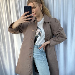 May include: A brown long-sleeved jacket with a button-up front and a white t-shirt with a graphic print underneath. The person is wearing blue jeans.