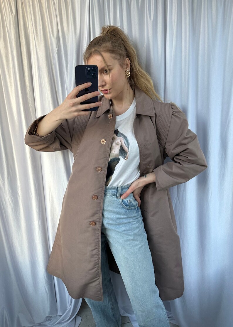 May include: A brown long-sleeved jacket with a button-up front and a white t-shirt with a graphic print underneath. The person is wearing blue jeans.