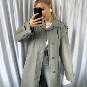 May include: A long, gray trench coat with a button closure and a belt at the waist. The coat has a classic design and is made of a lightweight fabric.