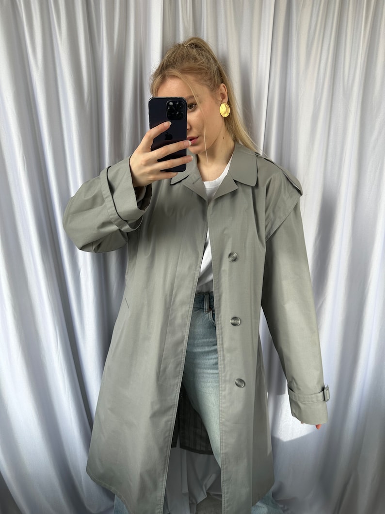 May include: A long, gray trench coat with a button closure and a belt at the waist. The coat has a classic design and is made of a lightweight fabric.