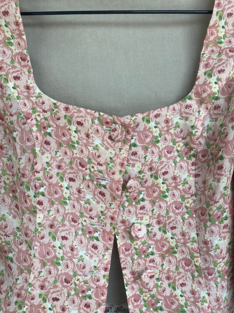 May include: Close-up of the front of a pink and green floral print top with a button closure. The top has a square neckline and a fitted bodice.