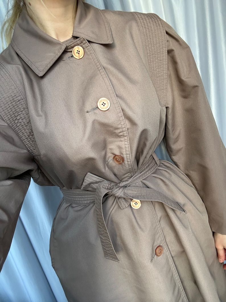 May include: A brown trench coat with a belt and buttons. The coat has a classic design and is made of a durable fabric.